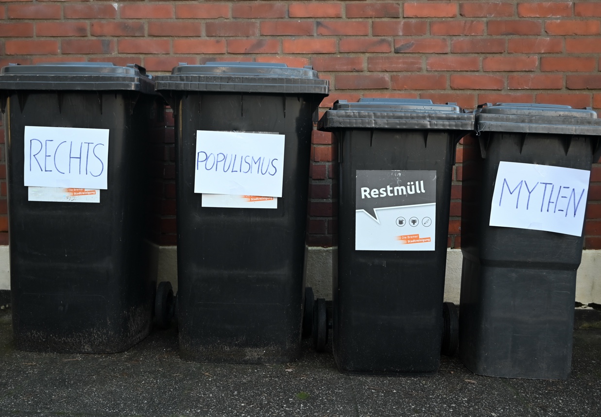 Vier schwarze Mülltonnen vor einer roten Ziegelmauer, beschriftet mit Zetteln, auf denen ‚RECHTS‘, ‚POPULISMUS‘, ‚RESTMÜLL‘ und ‚MYTHEN‘ steht. Die Beschriftung ‚Restmüll‘ auf einer der Tonnen stammt von der städischen Stadtreinigung, passend zum satirischen Kontext, in dem rechte Ideologien und Populismus symbolisch als Abfall dargestellt werden.